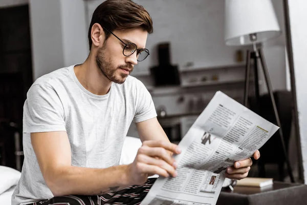 Young man in pajamas and eyeglasses sitting on bed and reading newspaper in the morning — Stock Photo