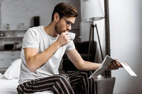 Young man in pajamas and eyeglasses drinking coffee and reading newspaper in the morning — Stock Photo