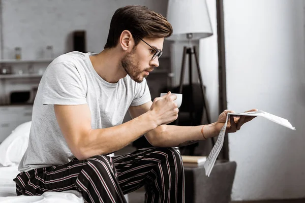 Joven guapo en pijama y anteojos sosteniendo taza de café y leyendo el periódico en la mañana - foto de stock