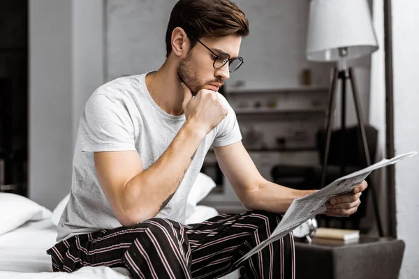 Beau jeune homme en pyjama et lunettes assis sur le lit et lire le journal le matin — Photo de stock