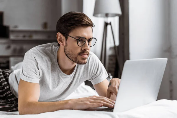 Handsome young man in eyeglasses and pajamas using laptop on bed in the morning — Stock Photo