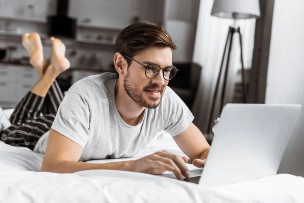 Улыбающийся молодой человек в очках и пижамах с ноутбуком на кровати — стоковое фото