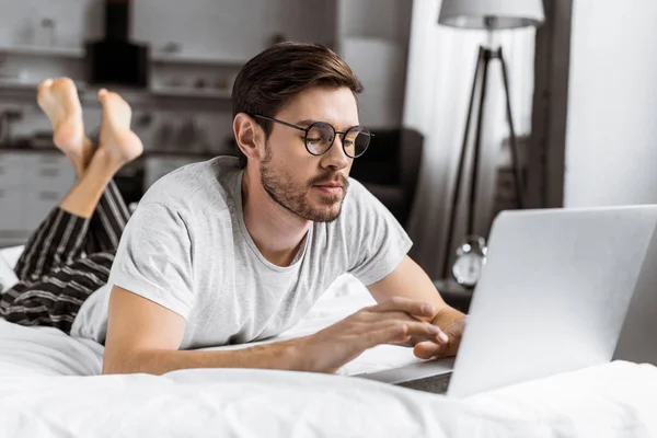 Bello giovane uomo in occhiali e pigiama con computer portatile sul letto — Foto stock