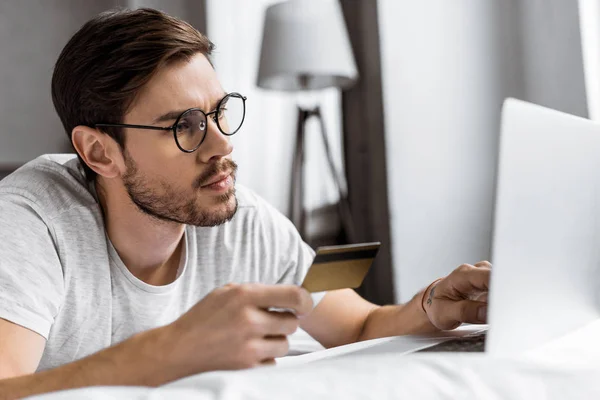 Bel giovane uomo con gli occhiali in possesso di carta di credito e utilizzando il computer portatile sul letto — Foto stock