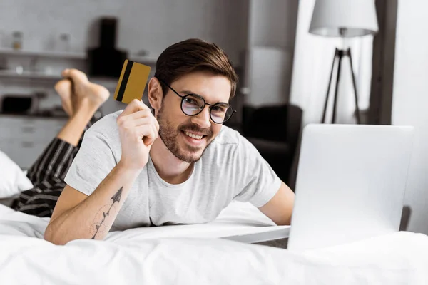 Schöner junger Mann mit Kreditkarte und lächelt in die Kamera, während er Laptop auf dem Bett benutzt — Stockfoto