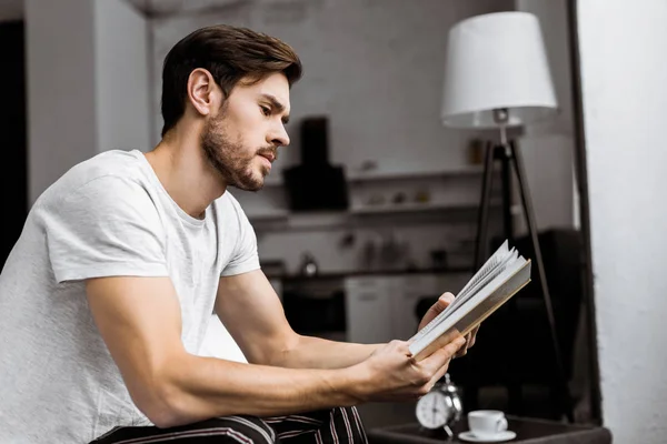 Vista lateral del joven guapo en pijama sentado y leyendo libro en casa - foto de stock