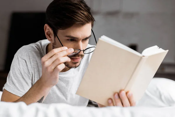 Красивый молодой человек регулирует очки и читает книгу, лежа на кровати — стоковое фото