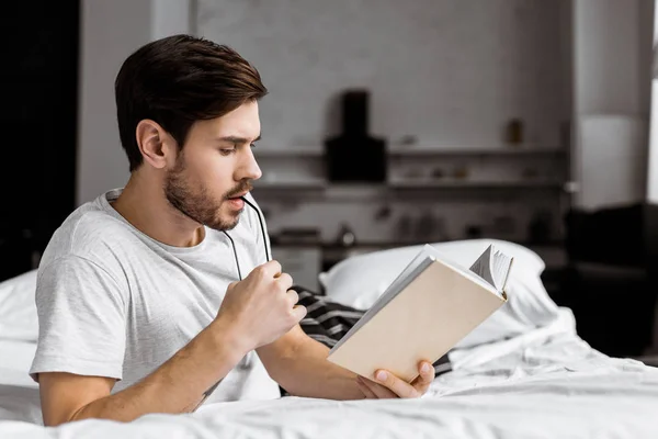 Joven guapo sosteniendo anteojos y leyendo libro mientras está acostado en la cama - foto de stock