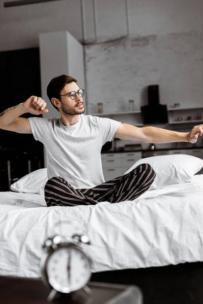 Jeune homme en pyjama et lunettes étirant tout en étant assis sur le lit le matin — Photo de stock