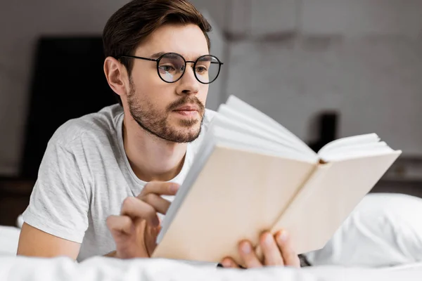 Beau jeune homme aux lunettes couché dans le lit et le livre de lecture — Photo de stock