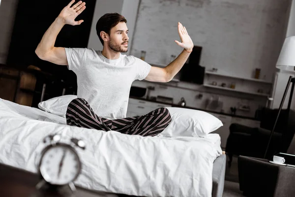 Handsome young man in pajamas sitting on bed and raising hands in the morning — Stock Photo