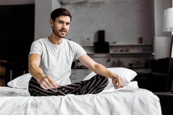 Handsome young man in pajamas sitting on bed and looking at camera — Stock Photo