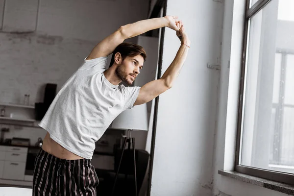 Handsome young man in pajamas stretching and looking at window in the morning — Stock Photo