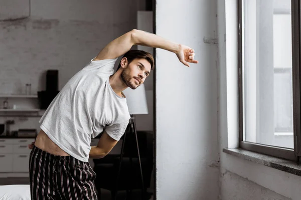 Handsome young man in pajamas exercising and looking at window in the morning — Stock Photo