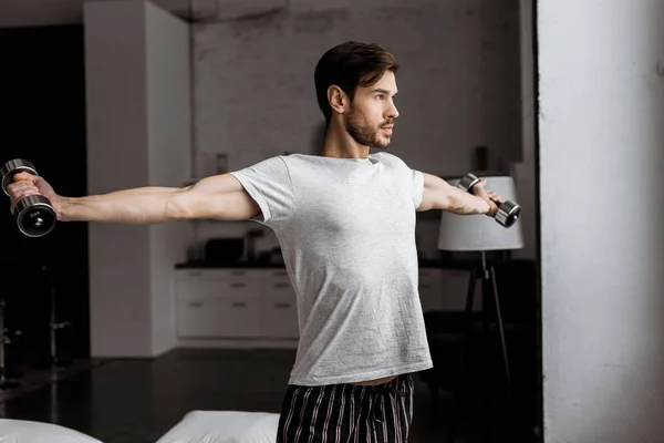 Schöner junger Mann im Pyjama, der mit Hanteln trainiert und zu Hause wegschaut — Stockfoto