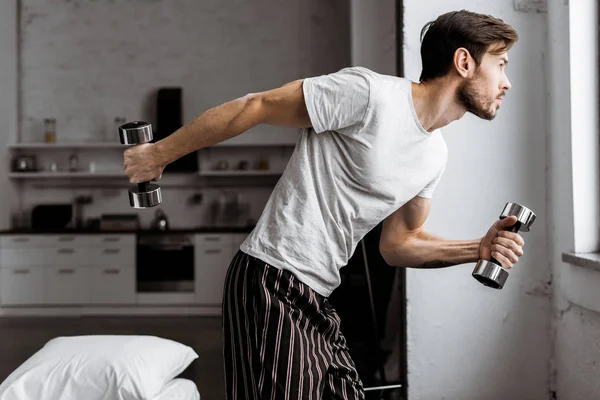 Joven en pijama sosteniendo mancuernas y mirando a la ventana en casa - foto de stock