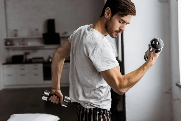 Beau jeune homme en pyjama faisant de l'exercice avec des haltères et regardant biceps à la maison — Photo de stock