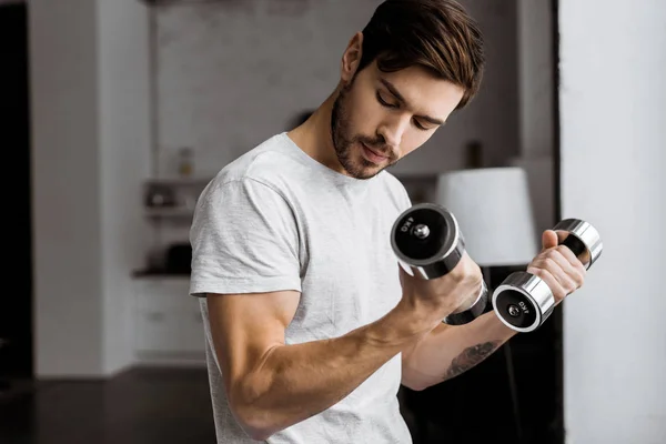 Schöner junger Mann trainiert mit Kurzhanteln und schaut zu Hause auf den Bizeps — Stockfoto