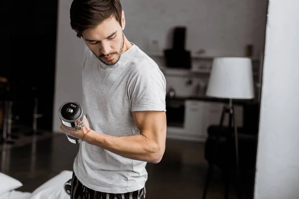 Beau jeune homme tenant des haltères et regardant biceps à la maison — Photo de stock