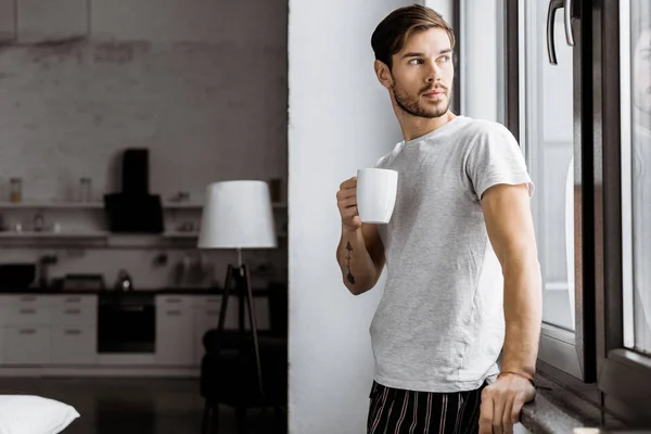 Jovem atraente em pijama com caneca de café inclinado para trás na janela em casa — Fotografia de Stock