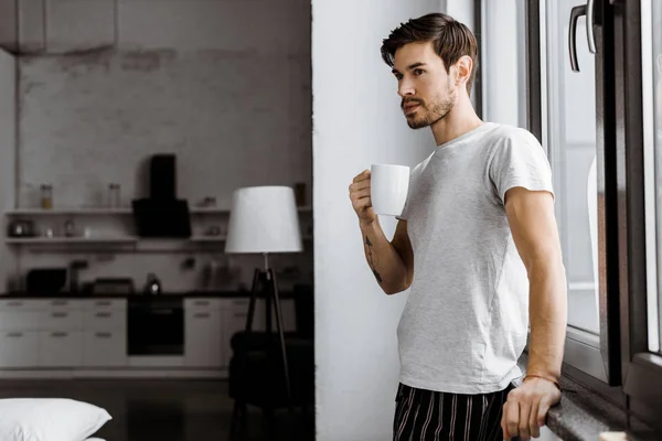 Beau jeune homme en pyjama avec tasse de café appuyé sur la fenêtre à la maison — Photo de stock