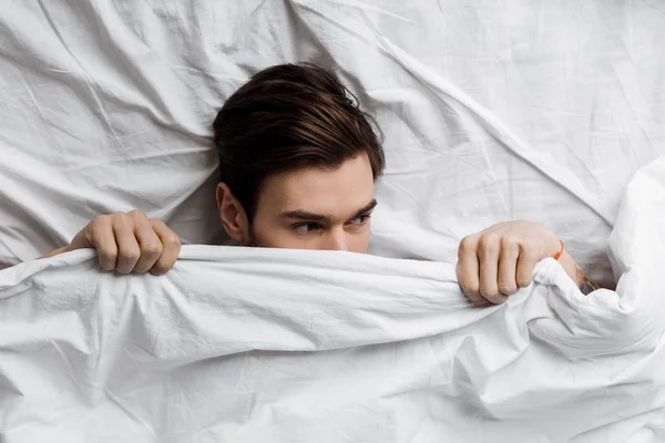 Von oben: schöner junger Mann versteckt sich zu Hause unter einer Decke im Bett und schaut weg — Stockfoto