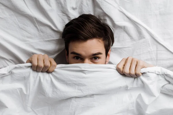 Vue de dessus du jeune homme se cachant sous la couverture dans le lit et regardant la caméra — Photo de stock