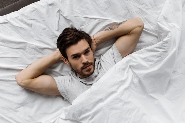 Вид сверху на привлекательного молодого человека, лежащего под одеялом в постели и смотрящего в камеру — стоковое фото