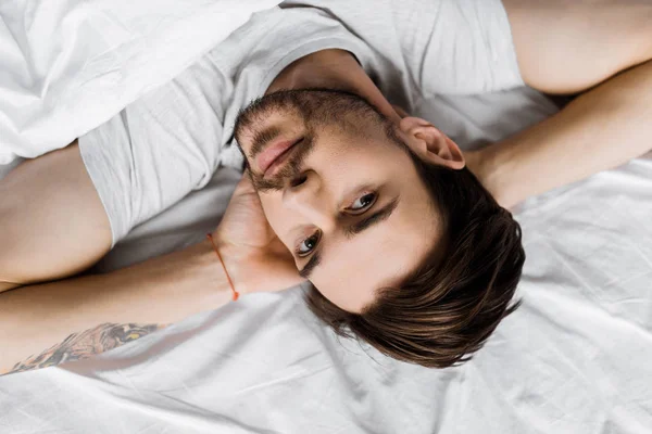 Top view of handsome young man lying under blanket in bed and looking at camera — Stock Photo