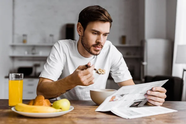 Joven guapo tomando cereal para el desayuno y leyendo el periódico en casa - foto de stock