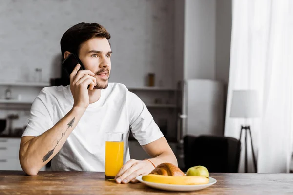 Joven guapo hablando por teléfono inteligente durante con un vaso de jugo de naranja y frutas en la mesa en casa - foto de stock