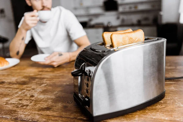 Schnappschuss eines jungen Mannes, der zu Hause Kaffee trinkt, mit Toaster im Vordergrund — Stockfoto