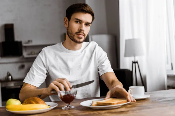 Schöner junger Mann, der zum Frühstück mit Marmelade und Früchten anstößt und zu Hause in die Kamera schaut — Stockfoto