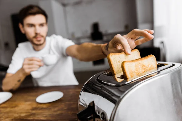 Close-up shot of young man drinking coffee and taking toast from toaster — Stock Photo