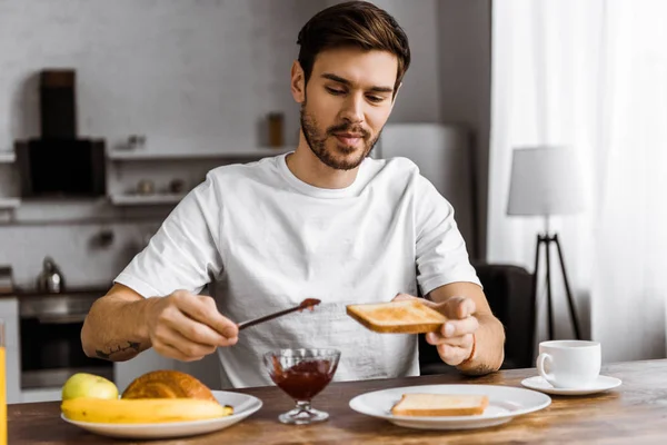 Усміхнений молодий чоловік наносить варення на тост у вихідні вранці вдома — стокове фото