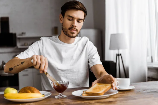 Atractivo joven aplicando mermelada en tostadas en fin de semana por la mañana en casa - foto de stock