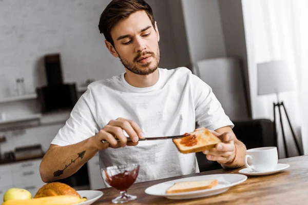 Handsome young man applying jam onto toast on weekend morning at home — Stock Photo