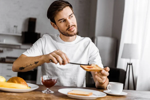 Schöner junger Mann, der zu Hause Marmelade auf Toast aufträgt — Stockfoto