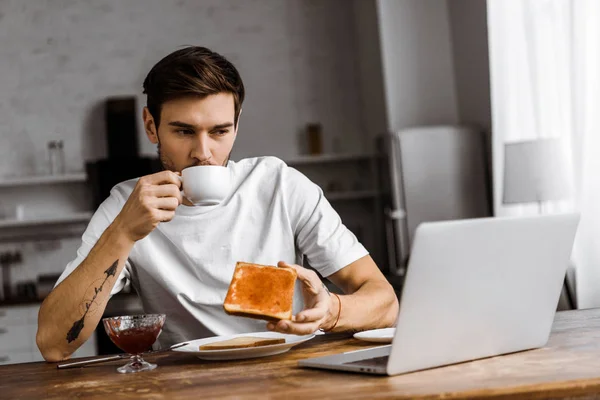 Handsome young freelancer eating toast with jam and coffee and looking at laptop screen at home — Stock Photo