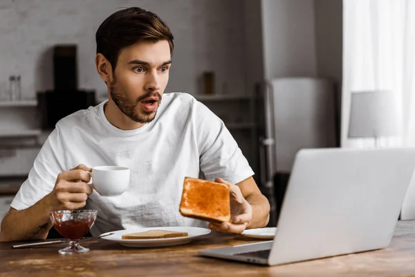 Шокированный молодой фрилансер ест тосты с джемом и смотрит на экран ноутбука дома — стоковое фото