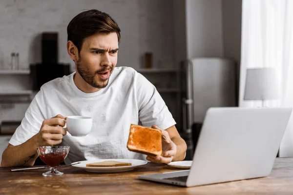 Confused young freelancer eating toast with jam and looking at laptop screen at home — Stock Photo