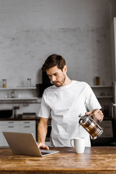 Jovem freelancer bonito com xícara de chá, croissant e laptop trabalhando na cozinha em casa — Fotografia de Stock