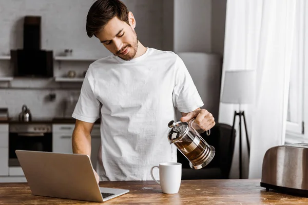 Atractivo joven freelancer con portátil verter té en la taza en la cocina en casa — Stock Photo
