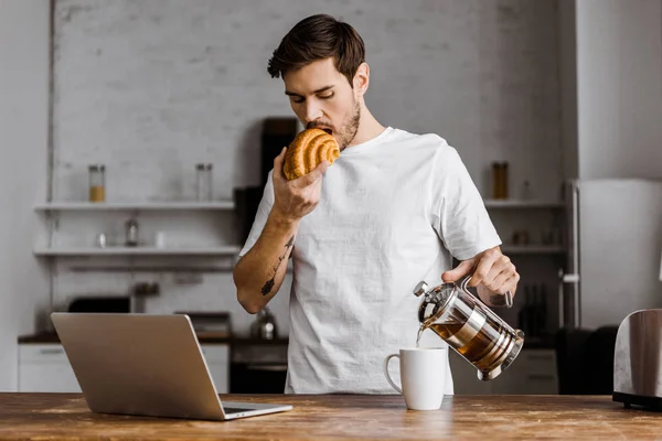 Atractivo joven freelancer con taza de té, croissant y portátil trabajando en la cocina en casa - foto de stock