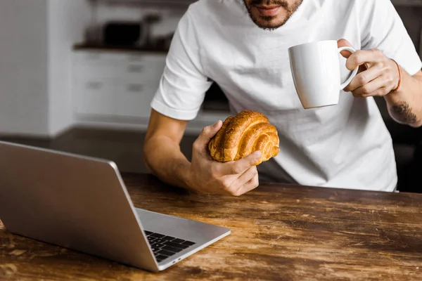 Tiro recortado de atraente jovem freelancer com xícara de chá, croissant e laptop trabalhando na cozinha em casa — Fotografia de Stock