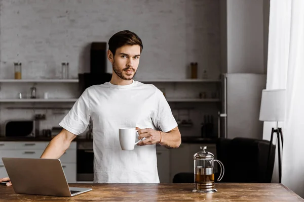 Jovem freelancer atraente com xícara de chá e laptop trabalhando na cozinha em casa — Stock Photo