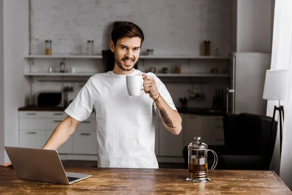 Jovem freelancer bonito com xícara de chá e laptop trabalhando na cozinha em casa — Stock Photo