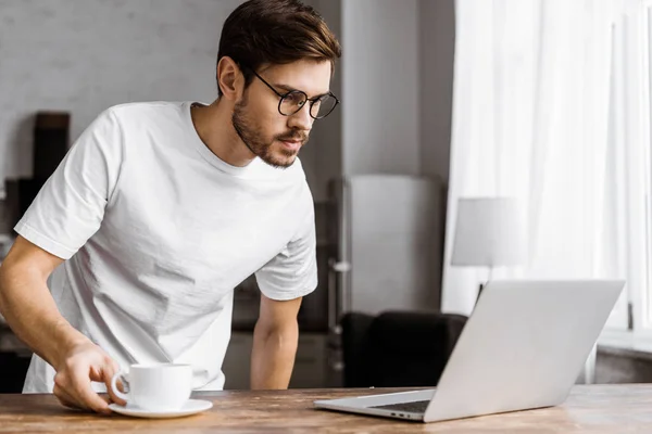 Handsome young freelancer in eyeglasses with coffee looking at laptop on kitchen at home — Stock Photo