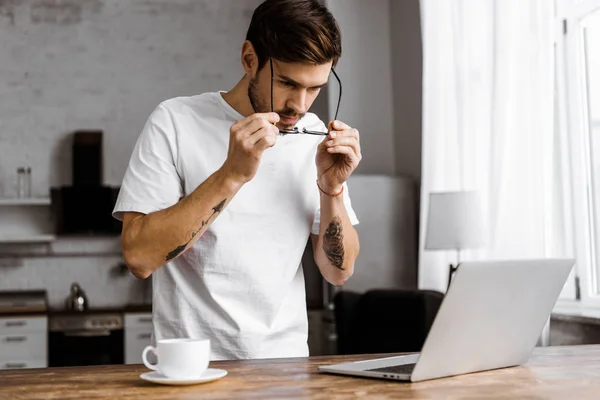 Hübsche junge Freiberuflerin in Brille mit Kaffee und Laptop in der heimischen Küche — Stockfoto