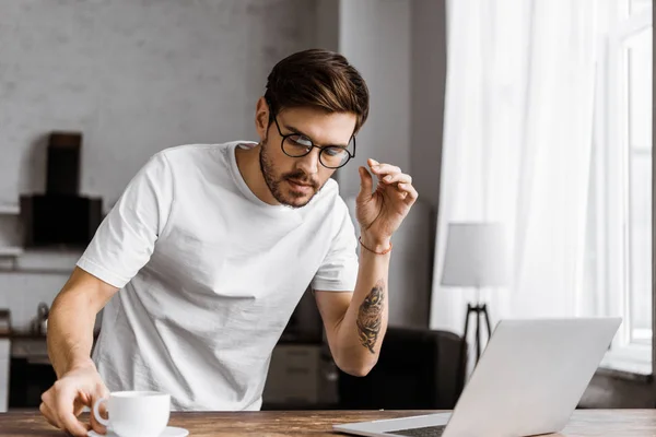 Handsome young freelancer with coffee and laptop on kitchen at home — Stock Photo